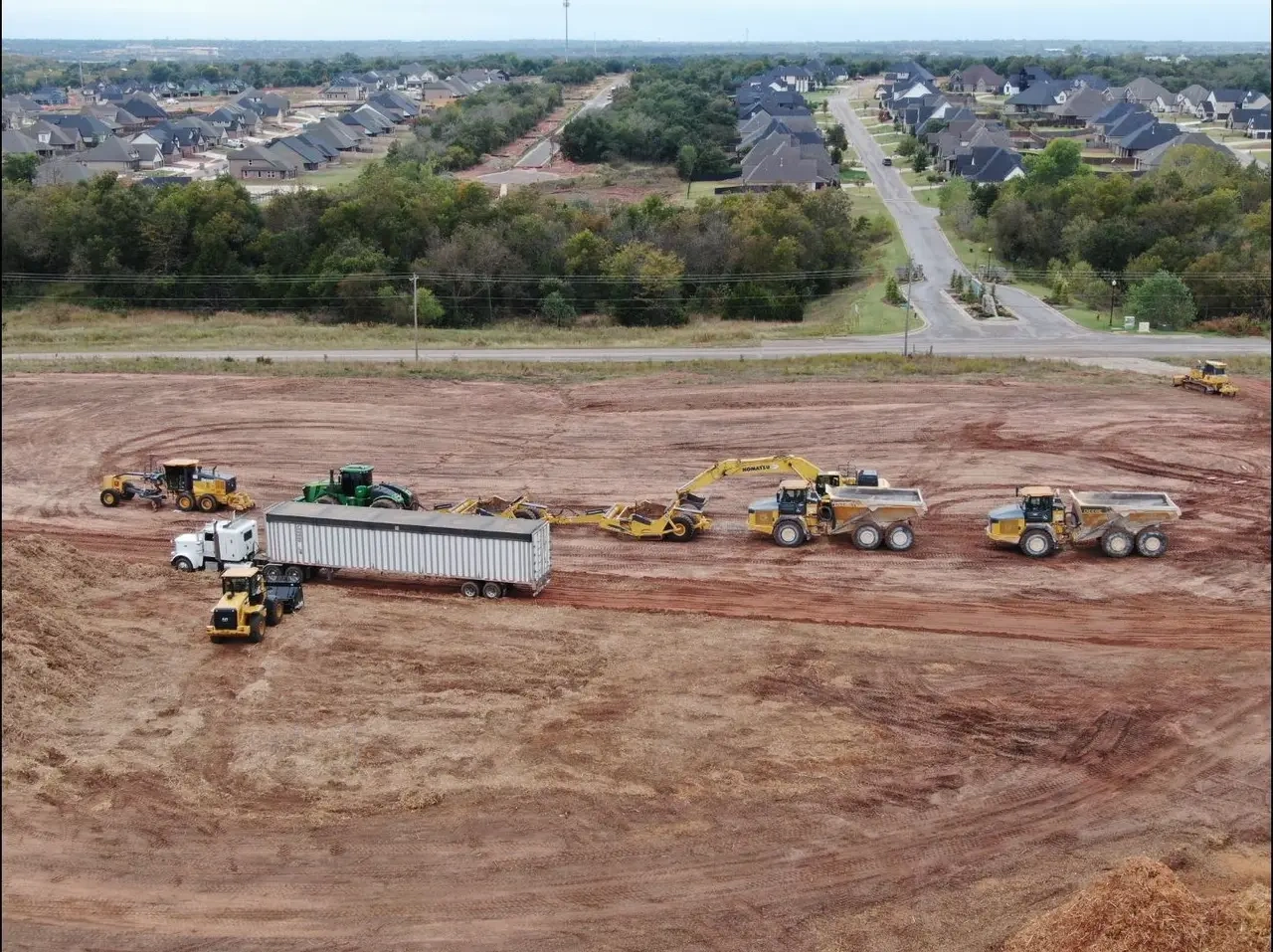 Aerial view of dozers, an excavator, articulated haul trucks, and a semi working a graded pad beside an Oklahoma subdivision
