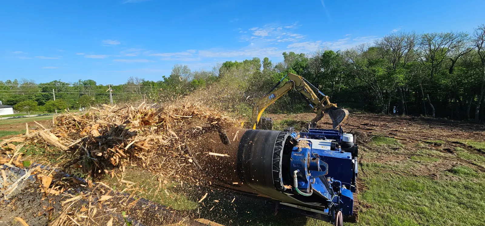Excavator feeding brush into a horizontal grinder that throws wood chips on an Oklahoma lot