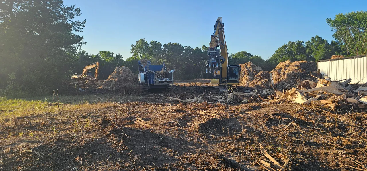 Excavators and a grinder cleaning up stumps and root wads on a cleared Oklahoma site at golden hour