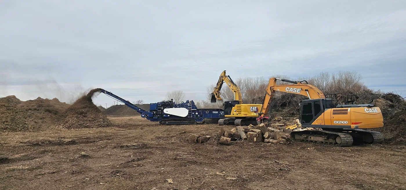 Horizontal grinder building a mulch pile while two excavators feed cleared timber in Oklahoma